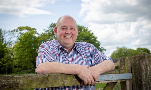 A man wearing a checked shirt leaning on a wooden gate smiling at camera 
